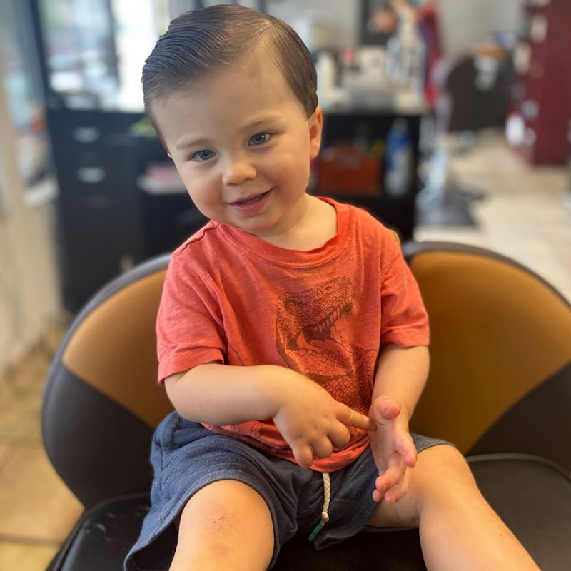 Young boy with a stylish haircut sitting in a barber chair, smiling and wearing a red t-shirt with a dinosaur graphic, in a barbershop setting.