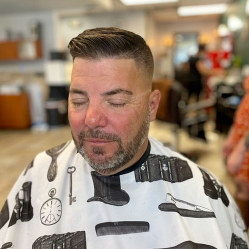Man with a stylish haircut and beard, wearing a barber cape with grooming tools pattern, sitting in a barbershop, relaxed atmosphere reflecting Joni's Gentleman's Cuts services.