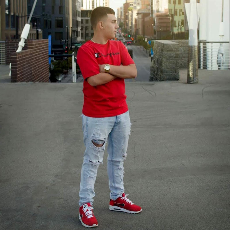 Young man standing confidently in a red shirt and distressed jeans, showcasing urban style in a city setting, embodying a relaxed yet fashionable atmosphere.