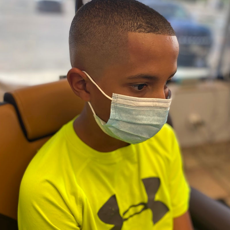 Young boy with a fresh buzz cut, wearing a blue mask and a bright yellow shirt, sitting in a barbershop chair, showcasing Joni's Gentleman's Cuts' commitment to stylish grooming and a relaxed atmosphere.