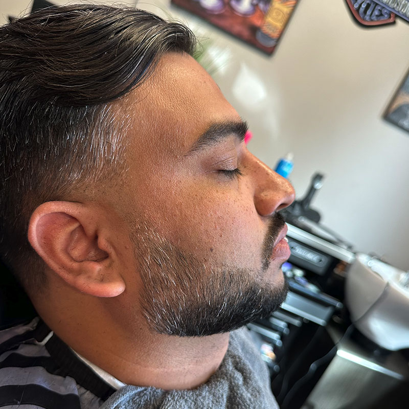 Man in barbershop chair receiving grooming service, showcasing a stylish haircut and well-defined beard, relaxed atmosphere with barber tools in background.