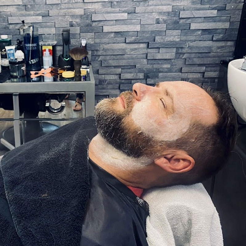 Man receiving a grooming service at a barber shop, relaxed with shaving cream on his face, surrounded by barber tools and products, emphasizing men's haircuts and grooming services.