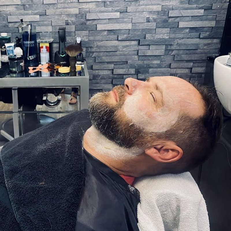 Man receiving a grooming service at a barber shop, relaxed with shaving cream on his face, surrounded by barber tools and products, emphasizing men's haircuts and grooming services.