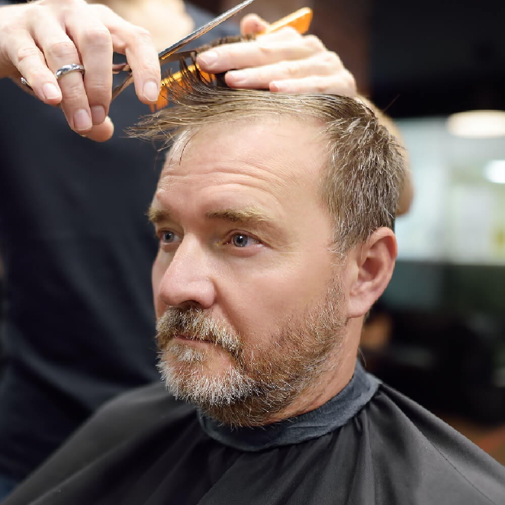 Older man with thinning hair and a gray-flecked beard getting a trim with scissors and comb.