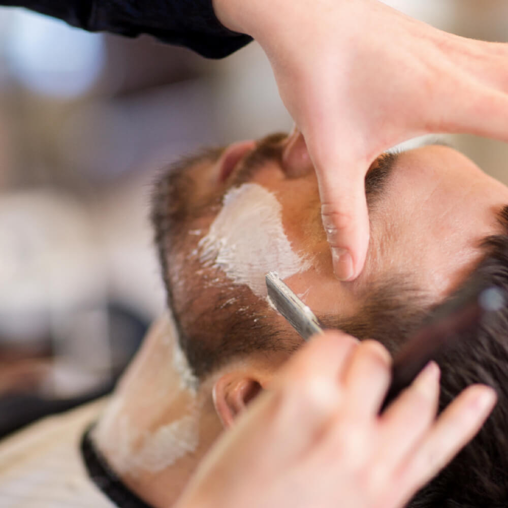 Barber using a straight razor to define a man's beard line along the cheek with shaving cream applied.