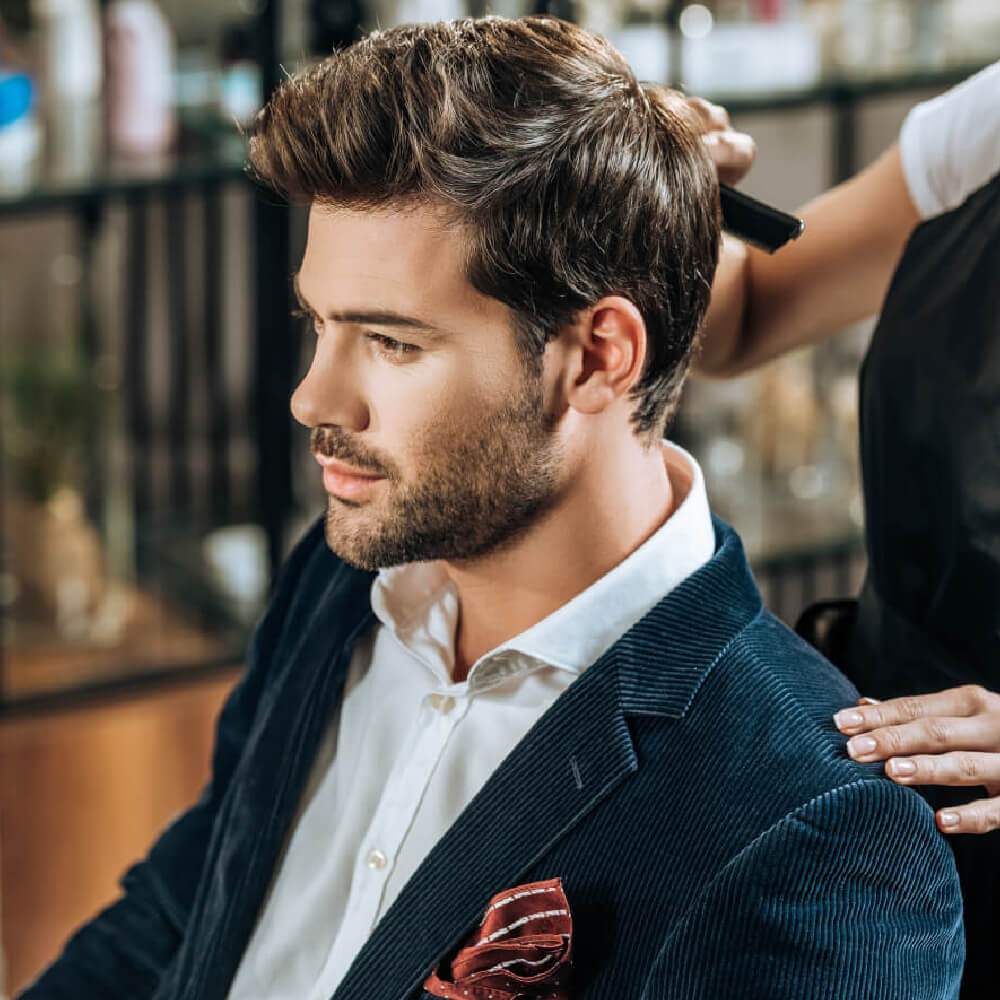 Man in a navy blazer and white shirt getting his voluminous, swept-back hair styled by a barber.