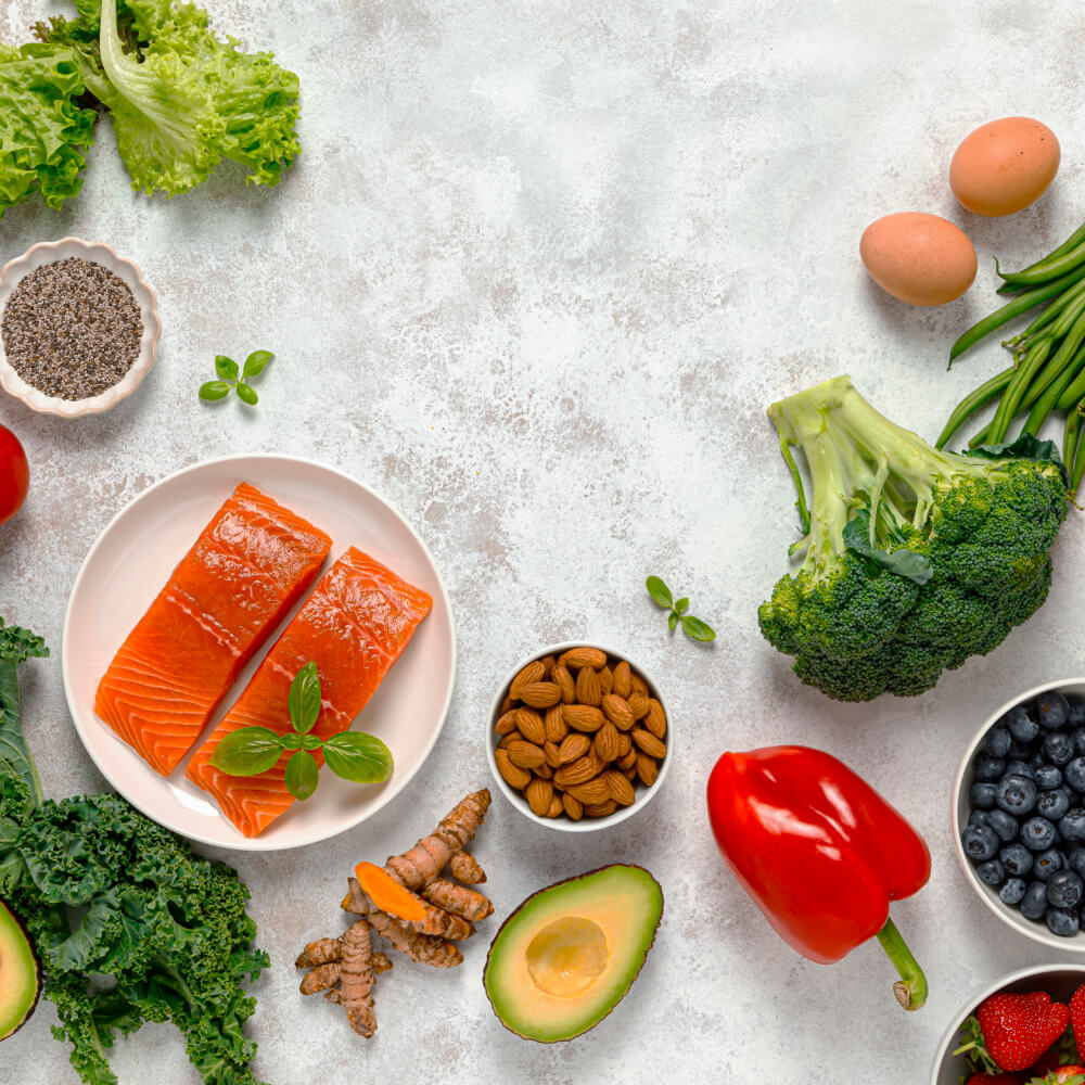 Flat lay of healthy foods including salmon, broccoli, kale, eggs, almonds, avocado, and berries.