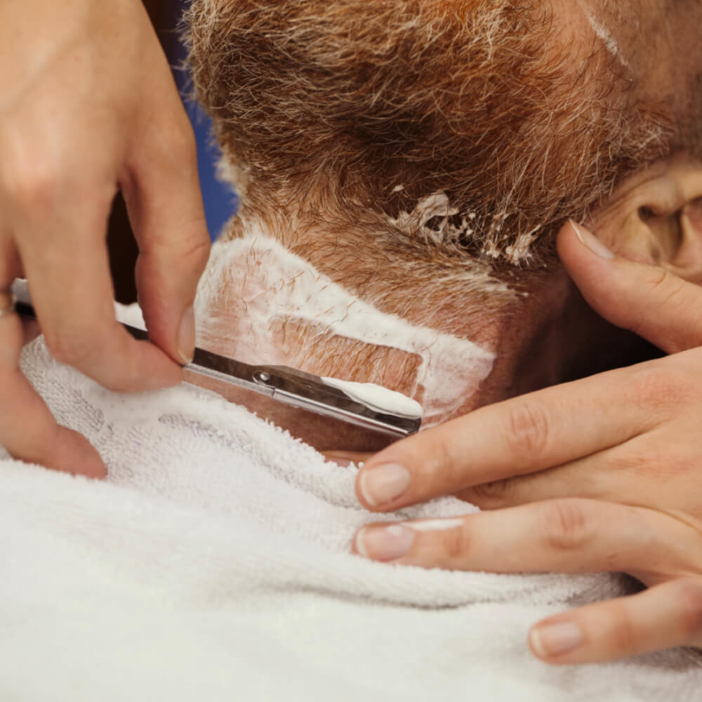 Close-up of a straight razor shaving the neckline below a man's red beard, edged with shaving cream.